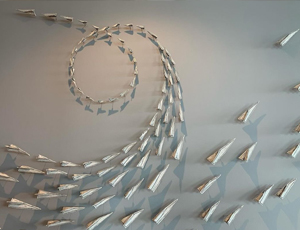 A photograph of a ceramic wall installation of a "flock" of paper airplanes by Jenifer Theom